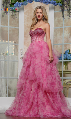 A woman stands indoors in front of mirrored doors with blue flowers, wearing the Strapless Print Prom Ball Gown: Colors Dress 3572, featuring a beaded corset bodice and a layered pink skirt.