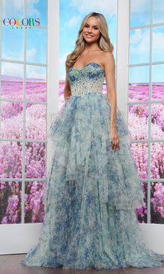 A woman indoors before glass doors reveals a purple flower field while wearing the Strapless Print Prom Ball Gown: Colors Dress 3572—an elegant pastel blue and green gown with floral print and a beaded corset-style bodice.