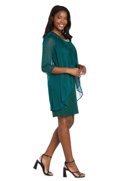 A woman stands sideways, smiling, in the R&M Richards 3594 Short Semi-Formal Jacket Dress—knee-length with sheer sleeves and overlay—paired with black heeled sandals.