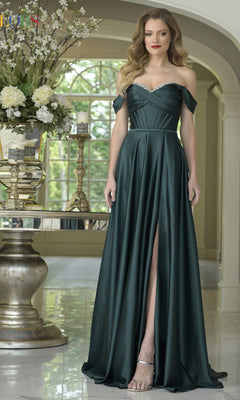 A woman in the Long Sweetheart Prom Dress: Colors Dress 3606, featuring a dark green off-the-shoulder design with high slit, stands indoors beside a table adorned with a large floral arrangement.
