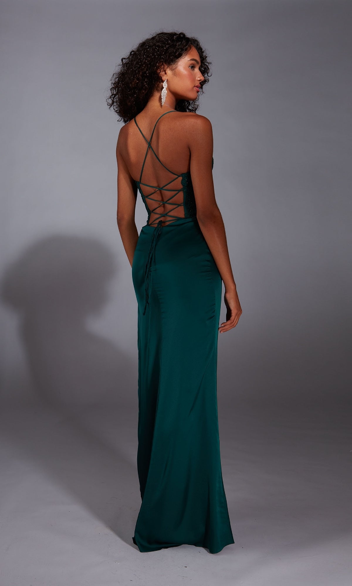 A woman in the Aerie Couture 362 long prom dress, featuring dark teal satin chiffon and a lace-up open back, stands facing away against a plain gray studio background.