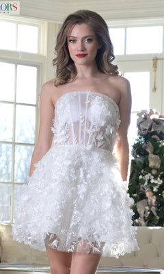 A woman wears the Strapless Floral Homecoming Dress: Colors Dress 3659, featuring 3-D flower embellishments, as she stands indoors by a large window and a decorated Christmas tree.