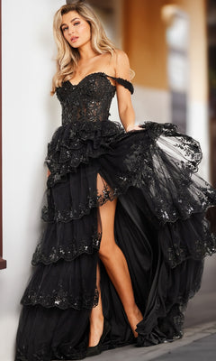 A woman poses in the Jovani 36687 long ruffled black prom ball gown, featuring a strapless off-shoulder design, layered details, lace and sequins, and a high slit.