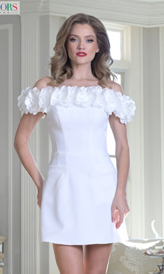 A woman stands indoors before large windows, wearing the Colors Dress 3679 Faux-Pocket Short Hoco Dress, which features a short off-the-shoulder design and delicate 3D floral embellishments on the neckline.
