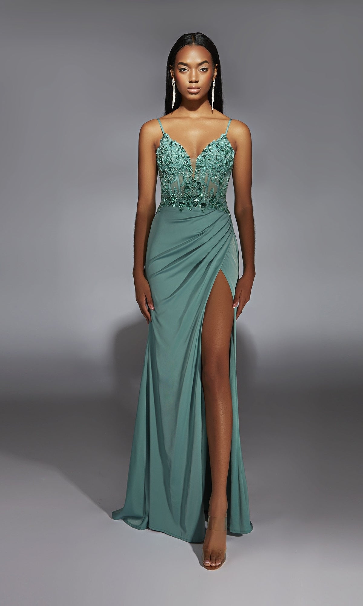 A woman in the Long Sage Green Prom Dress: Aerie Couture 369 with a lace bodice, spaghetti straps, and high slit pairs it with nude heels against a gray background.