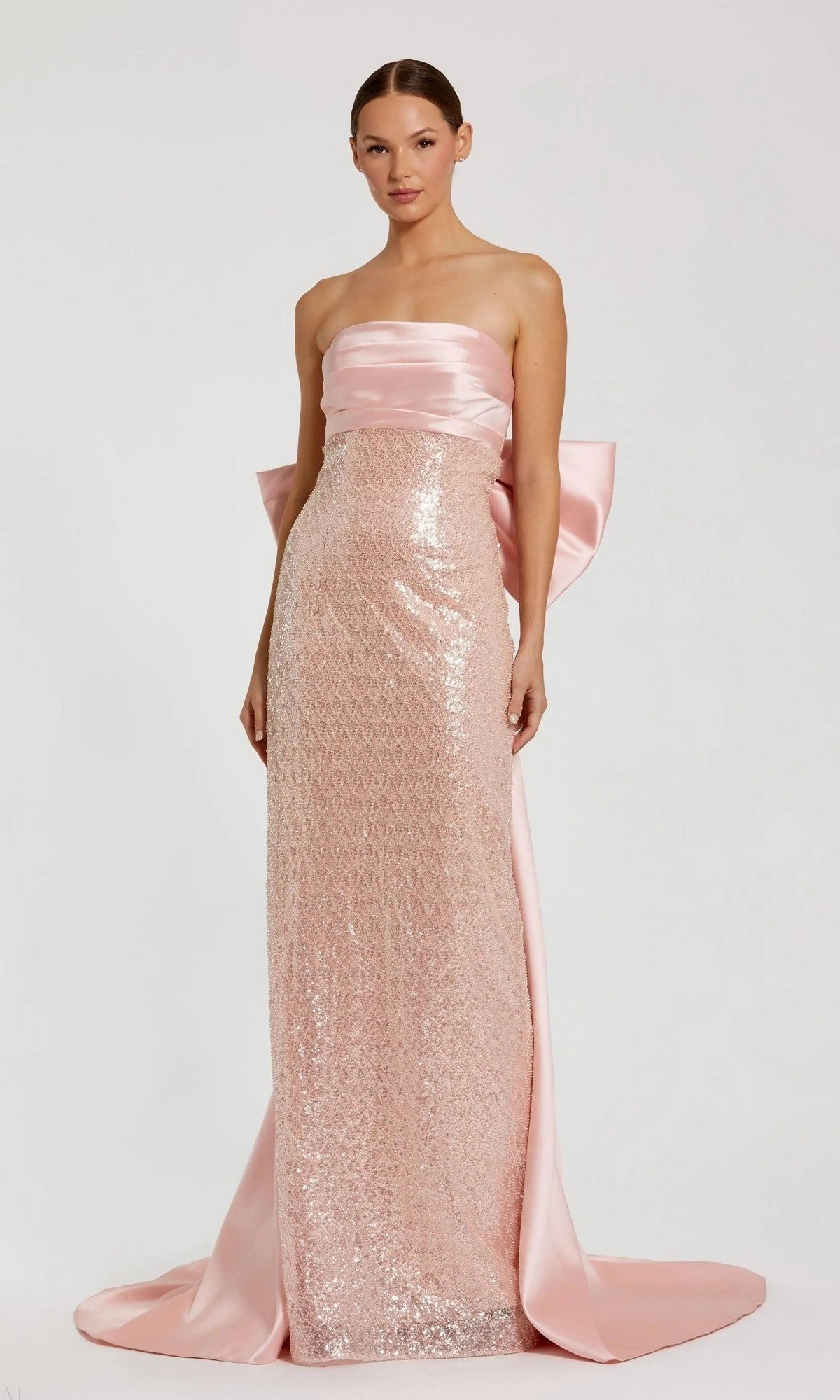 A woman models the Mac Duggal 37011 long formal dress in blush, featuring a strapless satin design with sequin details and a large bow at the back, set against a plain light background.