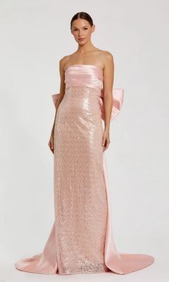 A woman models the Mac Duggal 37011 long formal dress in blush, featuring a strapless satin design with sequin details and a large bow at the back, set against a plain light background.