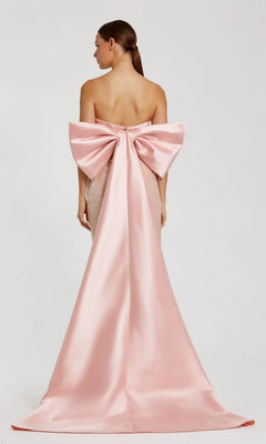 Woman wearing the Mac Duggal 37011 long formal dress, featuring a strapless satin design with a large bow and train at the back, standing against a plain light background.