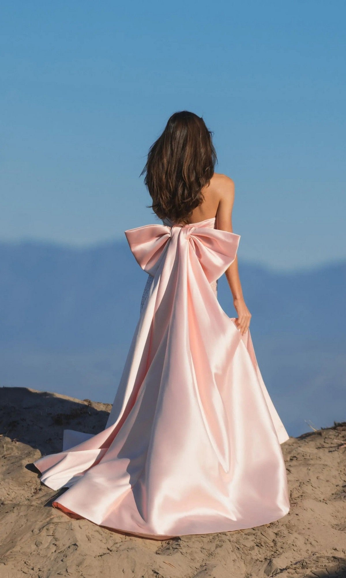 A woman with long brown hair stands outdoors, facing away from the camera in a strapless Mac Duggal 37011 satin gown featuring a large back bow, set against a clear blue sky.