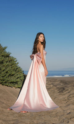 A woman stands on a sandy beach in the Mac Duggal 37011 long formal dress, a strapless satin gown featuring a large bow and train, with greenery and ocean in the background beneath a clear blue sky.