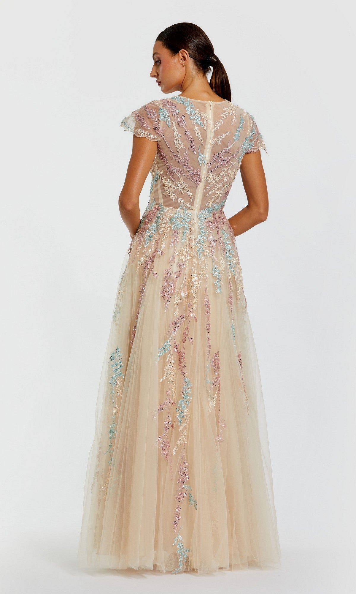 A woman showcases the back details and cap sleeves of the Mac Duggal 37014 long formal dress in nude, featuring pastel floral embroidery—an ideal choice for a mother-of-the-bride look against a simple backdrop.