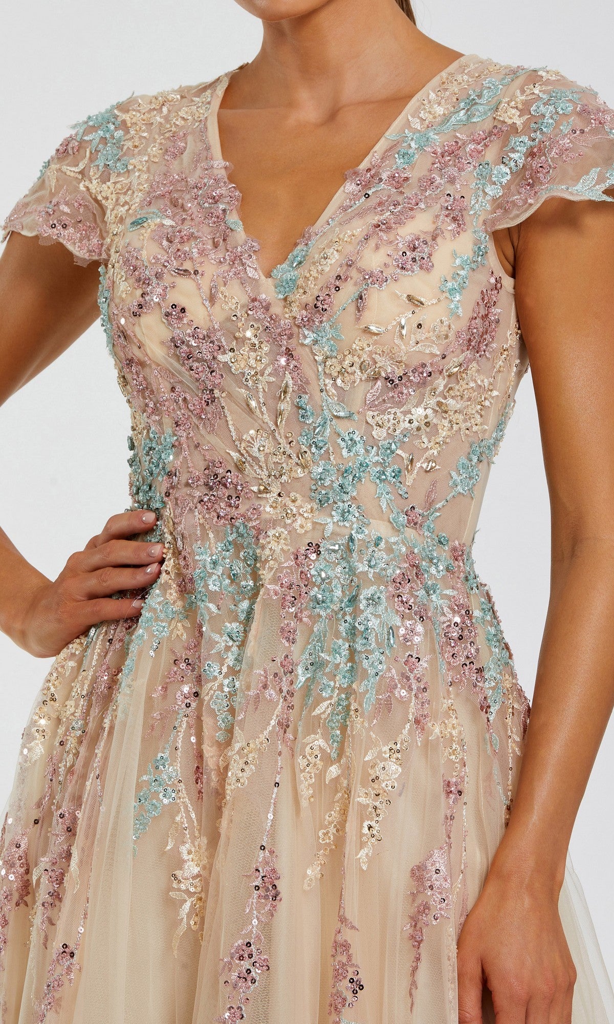 A woman in the Long Formal Dress: Mac Duggal 37014—a sheer, short-sleeved gown adorned with pastel pink, blue, and white floral sequins and embroidery on a beige base—makes a stunning statement for the modern mother-of-the-bride seeking a nude long formal dress with delicate detailing.