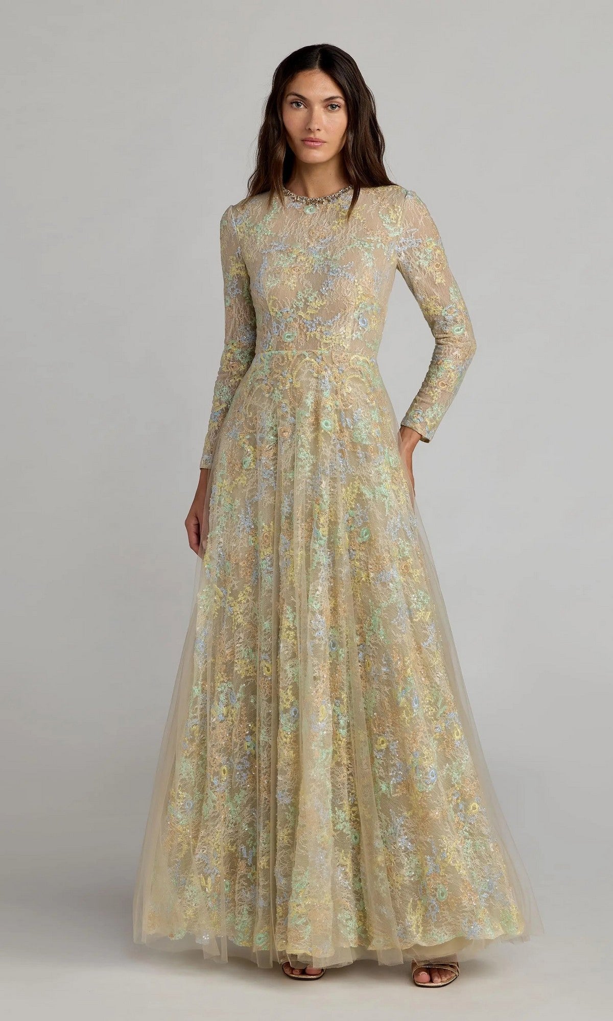 A woman wears the Mac Duggal 37016 long formal dress in Nude Multi, featuring long sleeves and a floor-length silhouette with a floral lace overlay, standing against a plain light background.