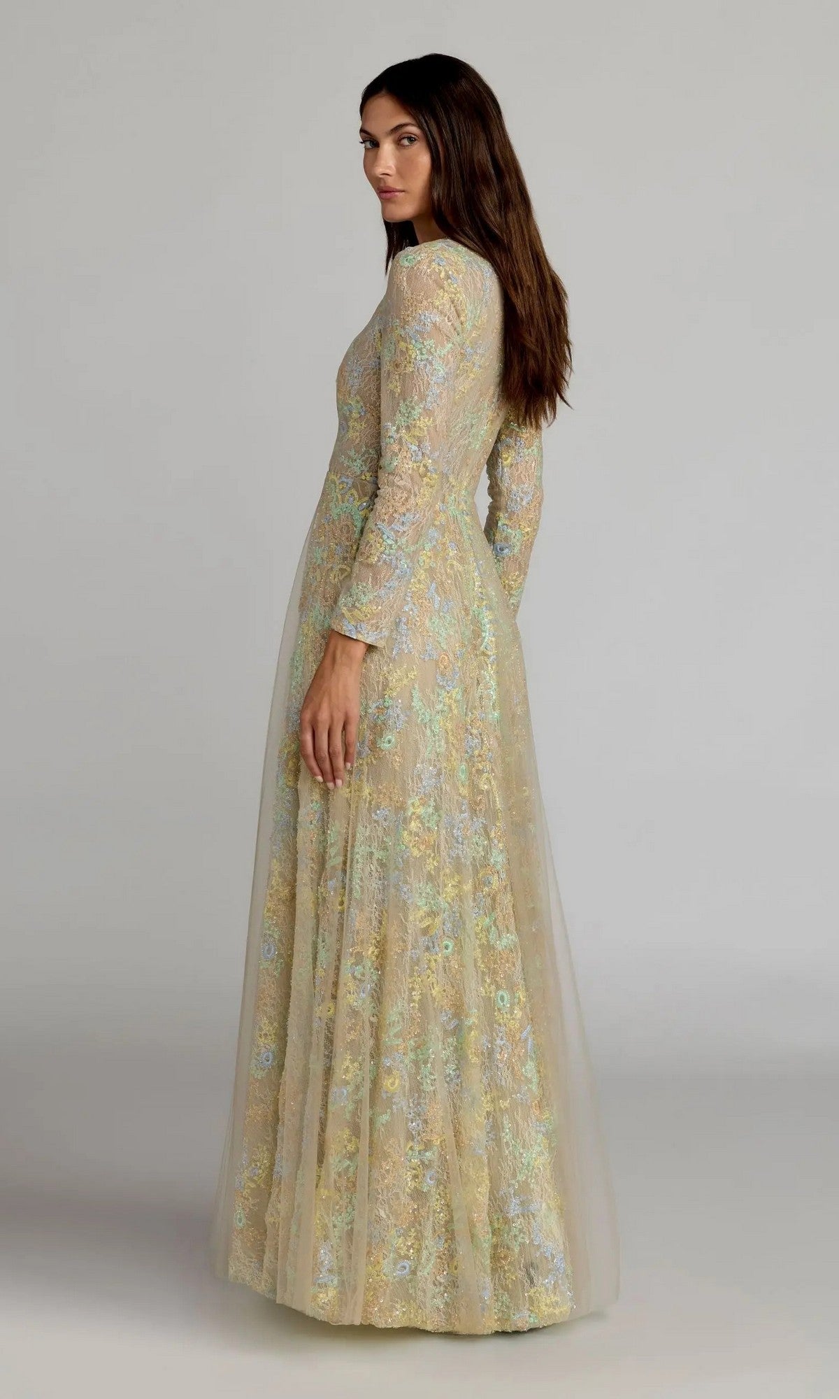 A woman wearing the Mac Duggal 37016 long formal dress—a modest, floor-length, pastel floral gown with long sleeves—stands against a plain background and looks over her shoulder.