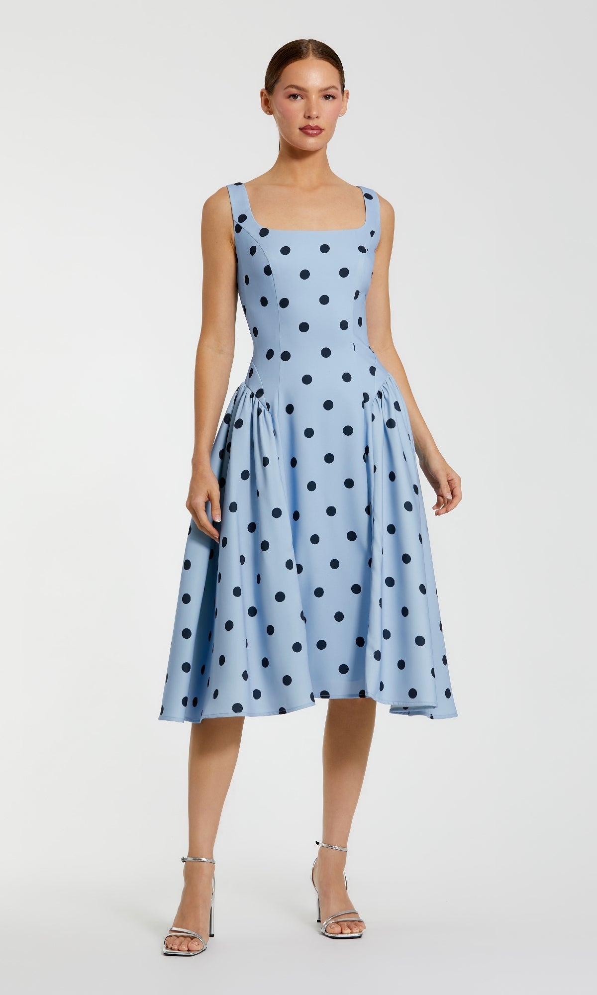 A woman in the Mac Duggal 37053 Polka-Dot Wedding-Guest Dress, a sky blue midi with black polka dots, and silver heels stands against a plain background.