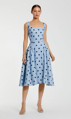 A woman in the Mac Duggal 37053 Polka-Dot Wedding-Guest Dress, a sky blue midi with black polka dots, and silver heels stands against a plain background.