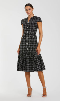A woman in the Mac Duggal 37071 semi-formal midi day dress, featuring black and white tweed with embellished buttons and a flared hem, pairs it with silver high-heeled sandals against a plain background.