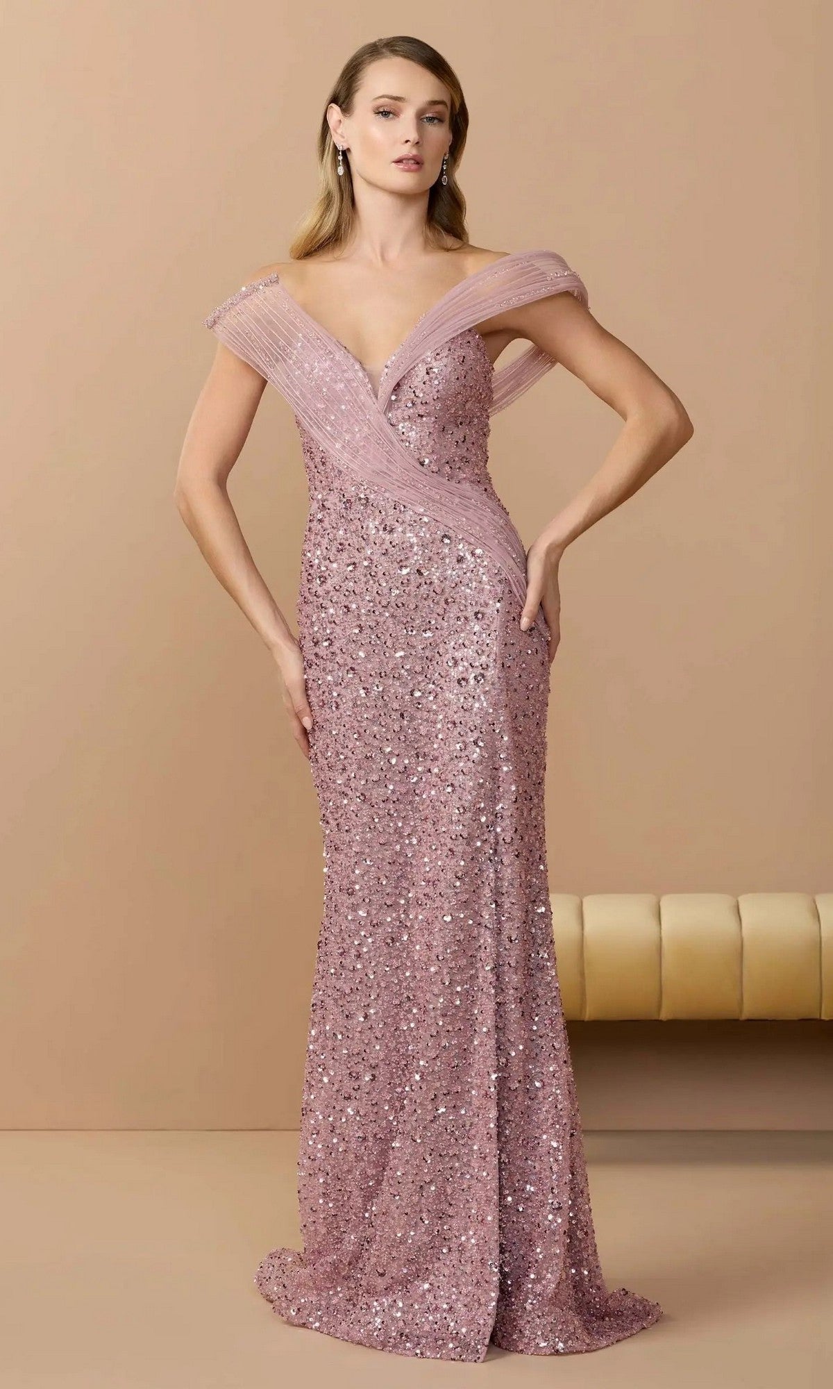 A woman stands against a beige background wearing the Mac Duggal 37085, a long formal, off-the-shoulder pink sequin couture evening gown.
