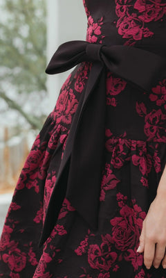 A person wears the Mac Duggal 37091 Floral Drop-Waist Party Dress, featuring a dark fabric with a red floral pattern and a large black bow at the waist.