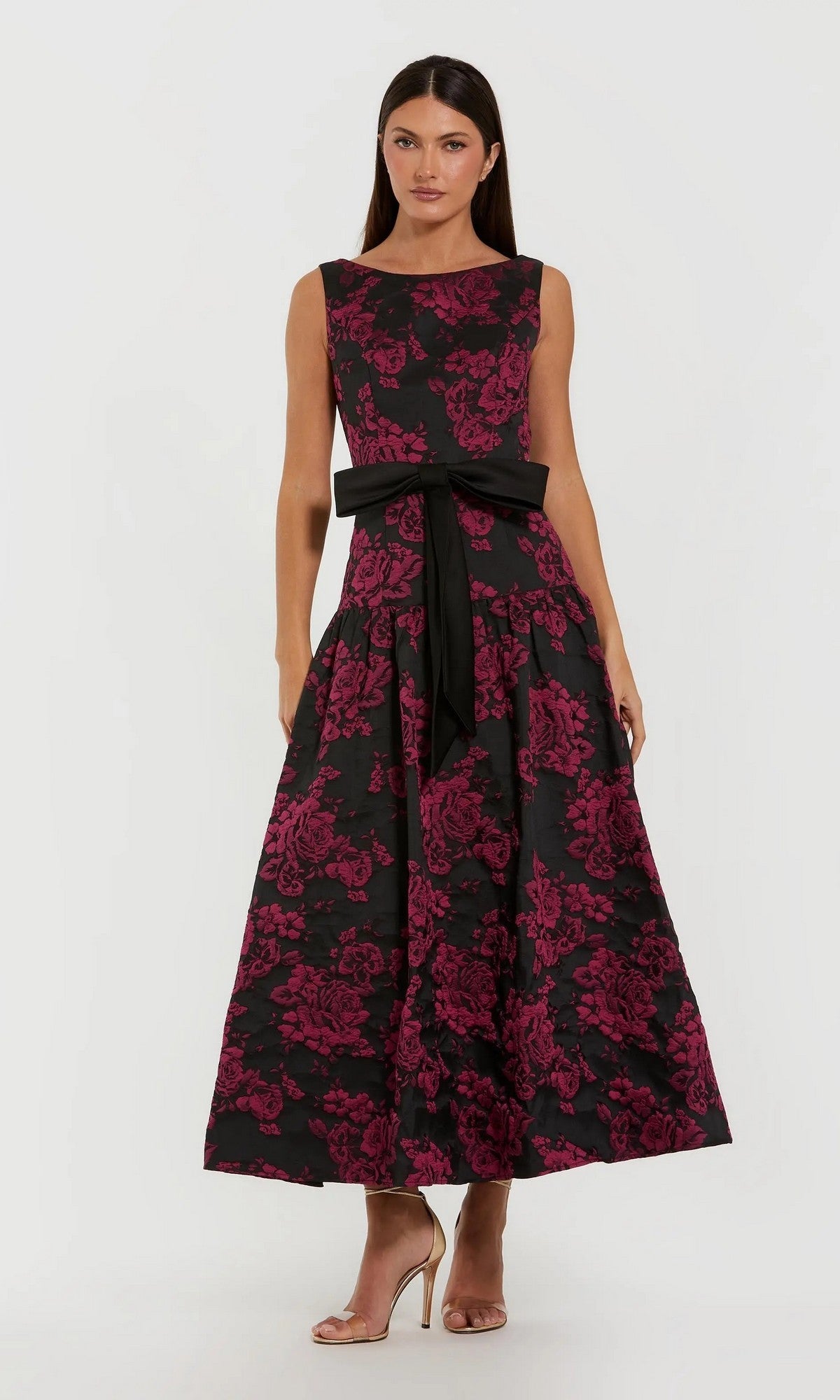 A woman stands against a plain background in the Mac Duggal 37091 Floral Drop-Waist Party Dress, a sleeveless ankle-length gown with a pink floral pattern, black waist sash, and beige high-heeled sandals—an elegant brocade look.