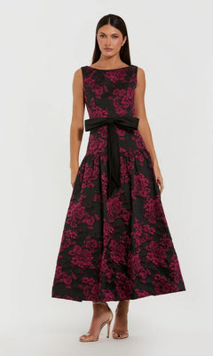 A woman stands against a plain background in the Mac Duggal 37091 Floral Drop-Waist Party Dress, a sleeveless ankle-length gown with a pink floral pattern, black waist sash, and beige high-heeled sandals—an elegant brocade look.