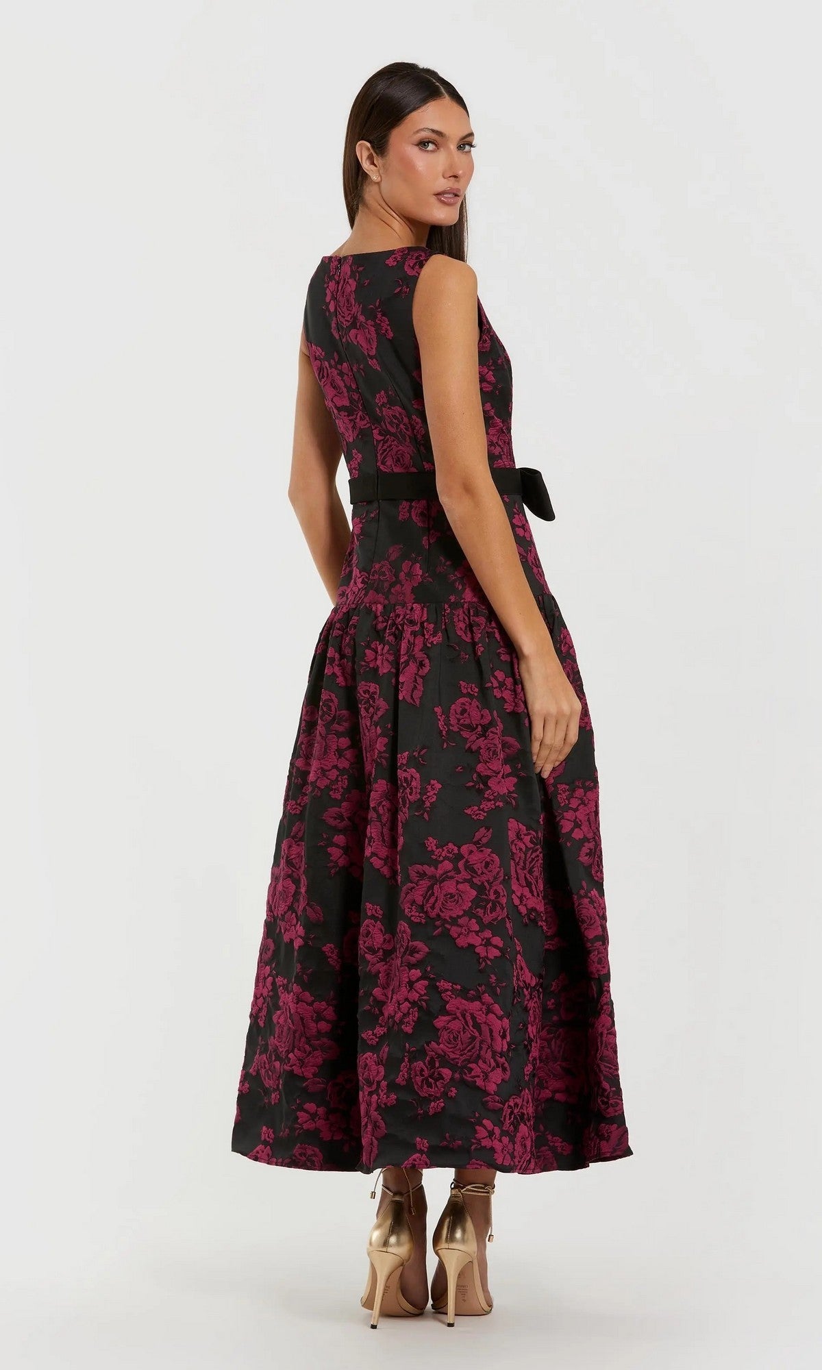 Woman wearing the Mac Duggal 37091 Floral Drop-Waist Party Dress—sleeveless, black with a pink floral pattern—paired with metallic heels, standing and glancing over her shoulder.