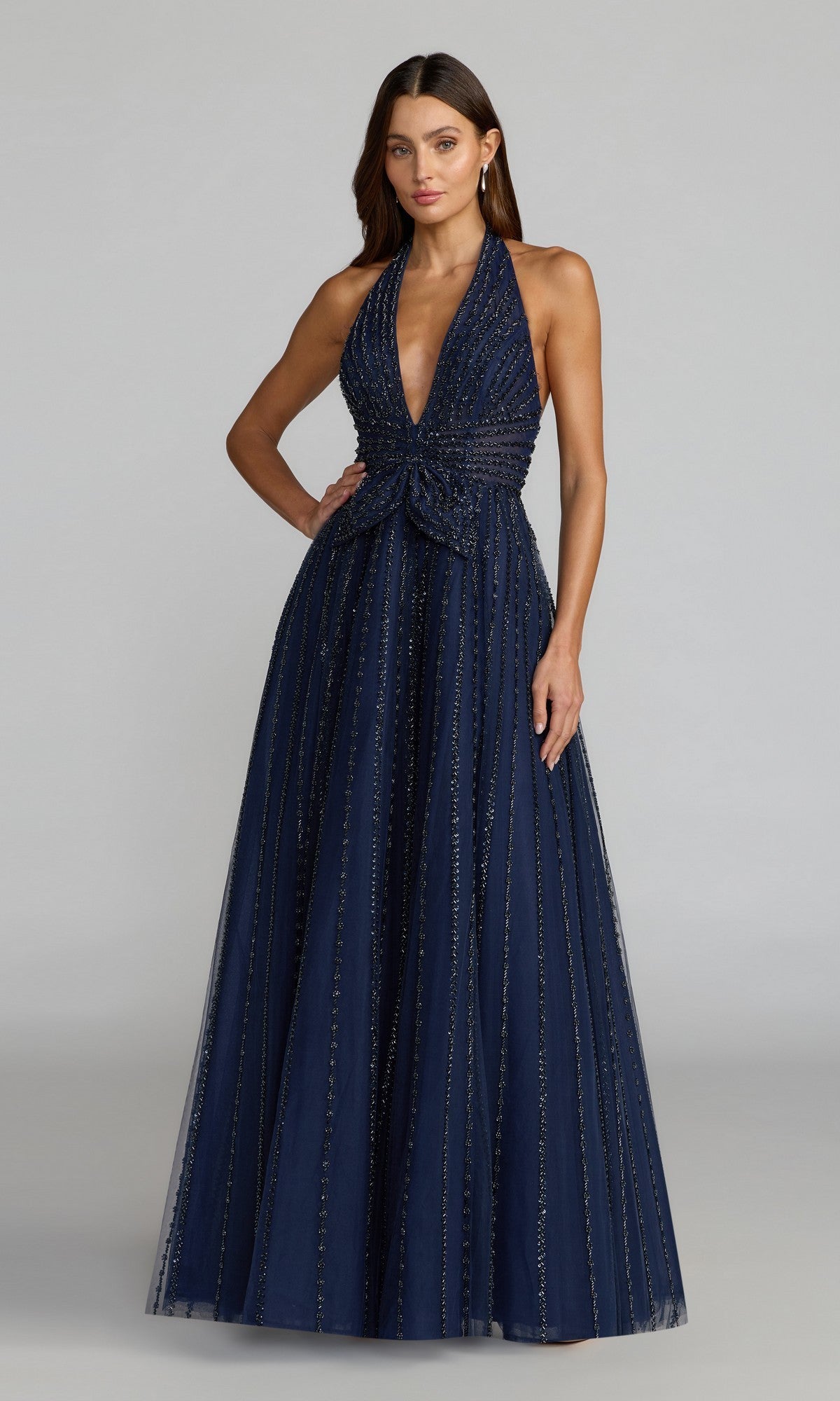 A woman stands against a plain light gray background wearing the Mac Duggal 37120 long formal dress in midnight—a sleeveless halter gown featuring a fitted bodice, bow detail at the waist, and vertical sparkling stripes.