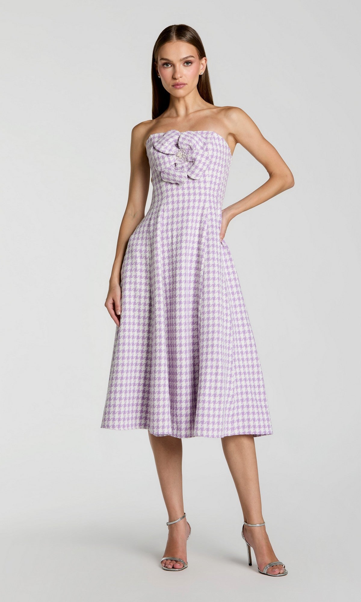 A woman stands against a plain background wearing the Strapless Gingham Midi Dress by Mac Duggal (37135) in lilac, featuring a strapless bodice with bow detail, paired with silver high-heeled sandals.