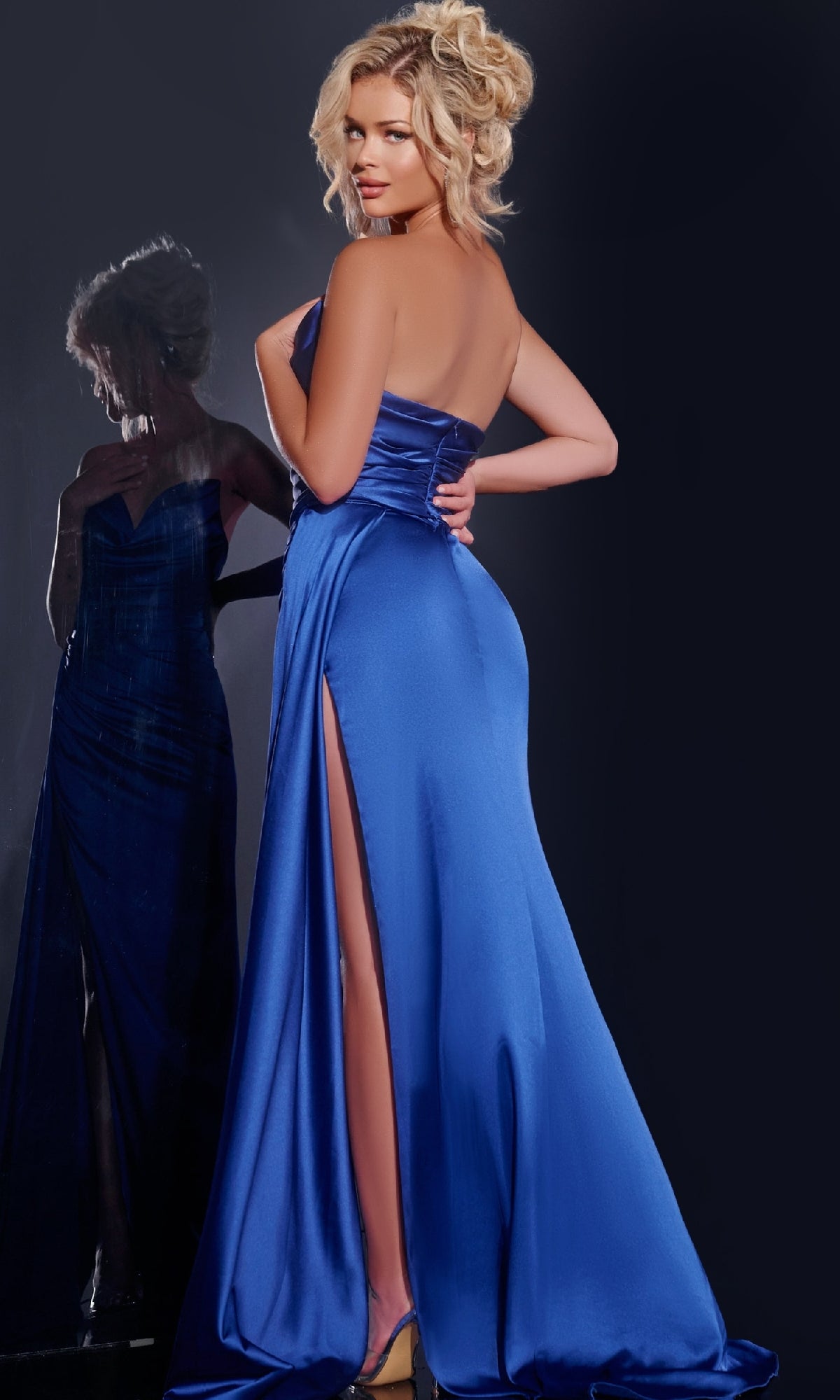 A woman poses with her back to the camera in the Jovani 37150 Long Prom Dress, a royal blue strapless satin gown with a high slit, its shimmery reflection visible in the background.