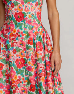 A person wearing the Floral Midi Wedding Guest Dress: Mac Duggal 37155, featuring a vibrant red, pink, and green floral print in an A-line midi silhouette, stands against a plain light gray background.