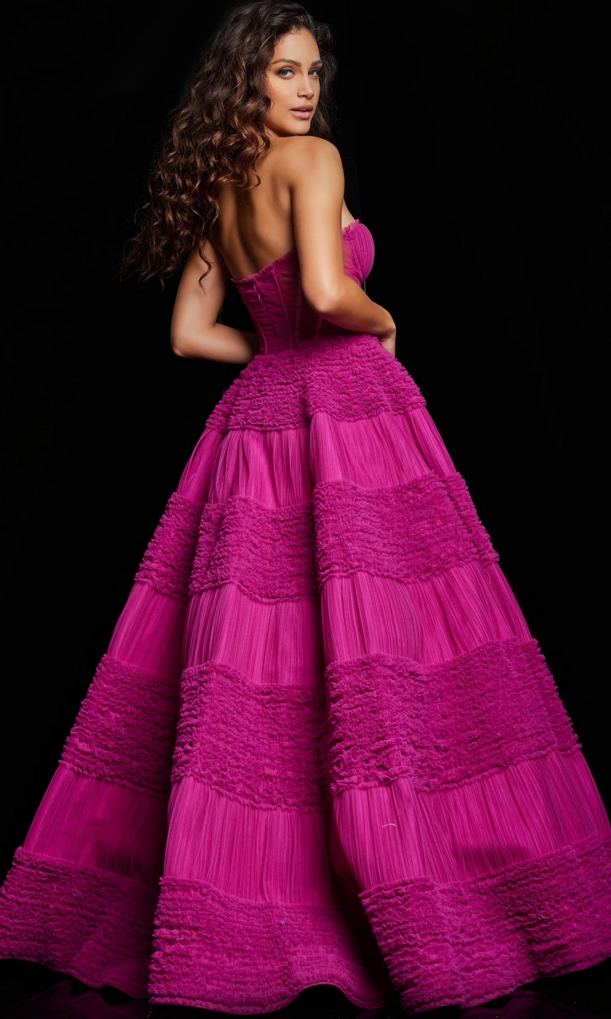 A woman with long wavy hair wears the Jovani 37157 Strapless Sheer-Corset Ball Gown in bright fuchsia with tiered, textured layers, standing against a black background.