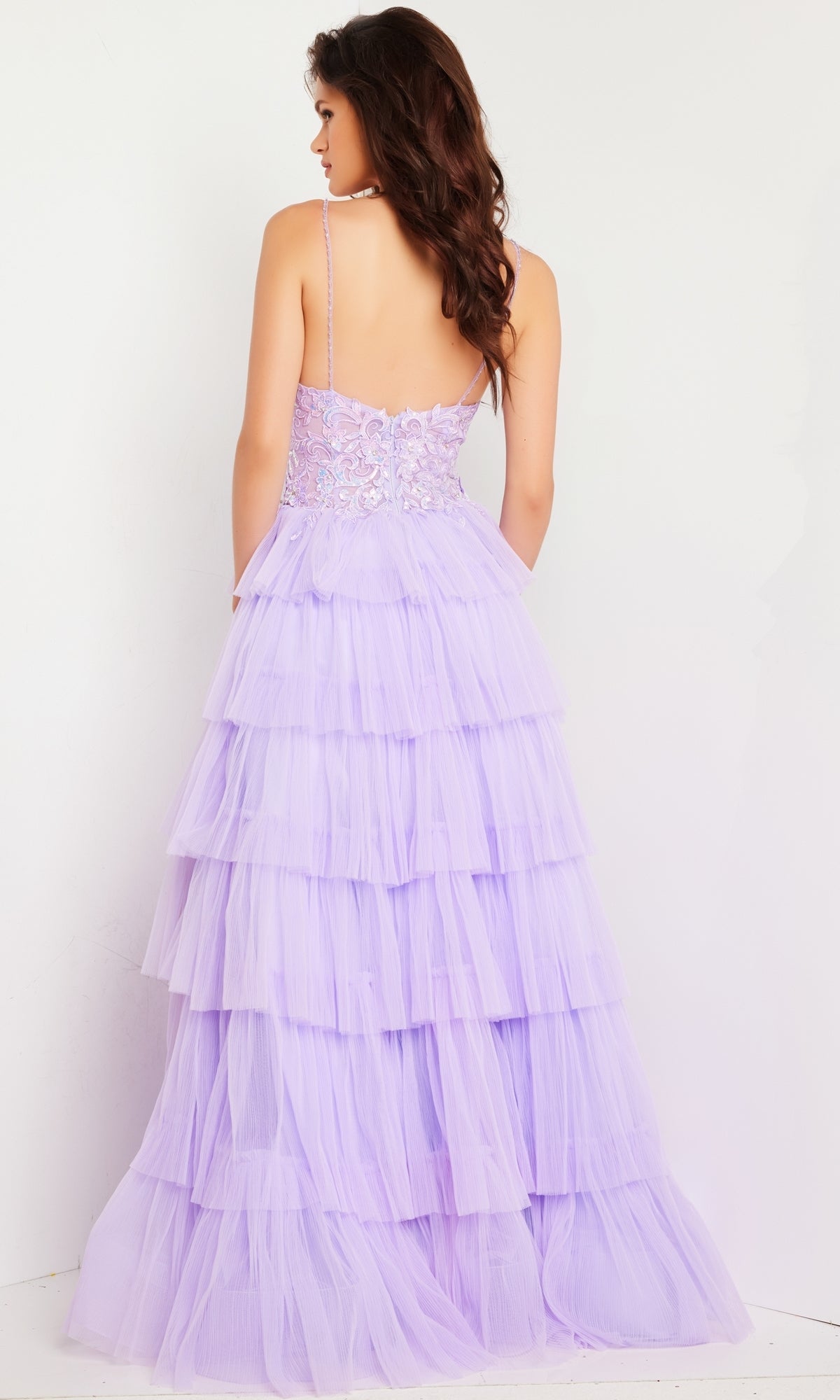 A woman faces away, wearing the Long Prom Dress 37190 by Jovani—a lavender ruffled tulle ball gown with a fitted, lace-embellished bodice and spaghetti straps—standing against a plain white background.