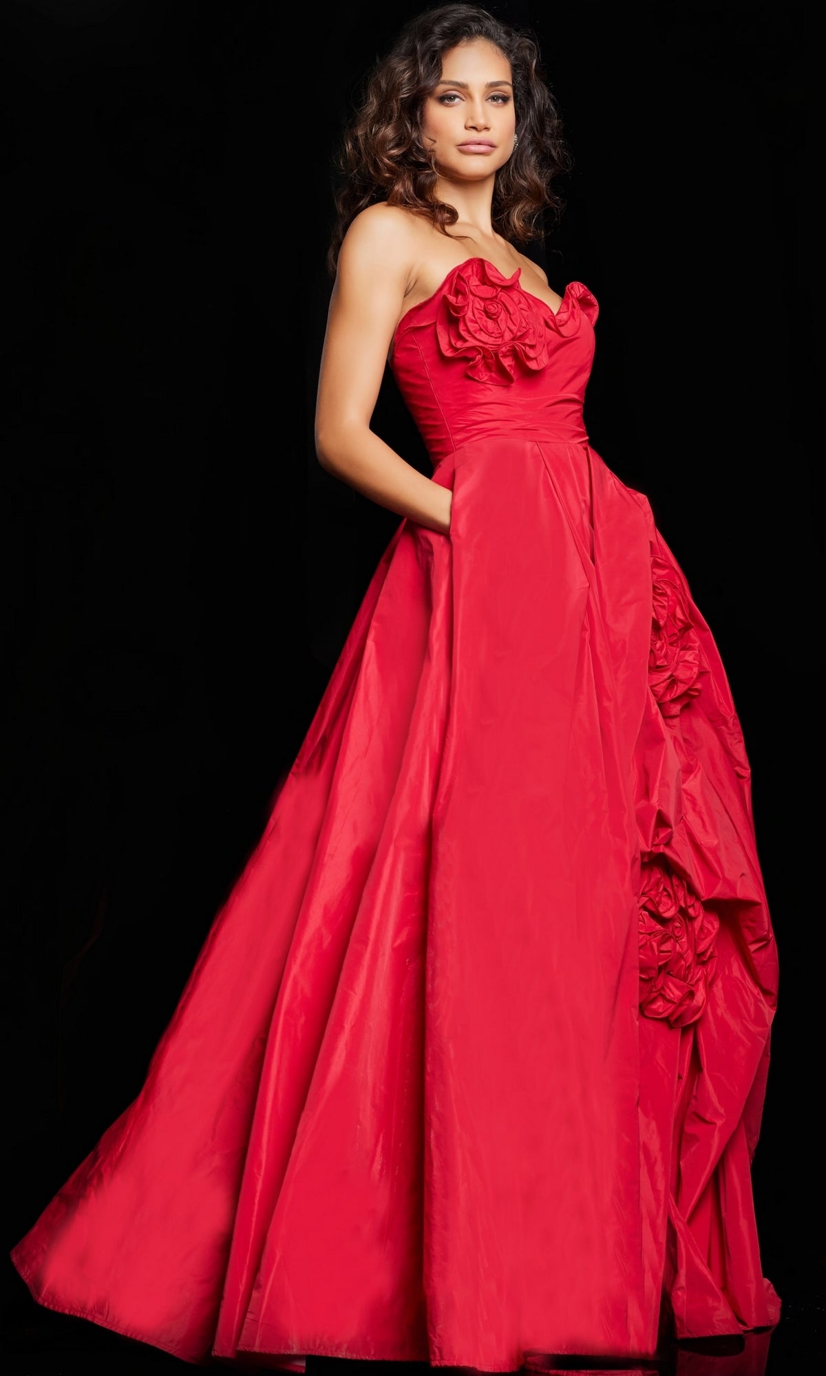 A woman stands against a black background in Jovani's Long Prom Dress 37266, a strapless, floor-length red ball gown featuring floral details and side pockets.