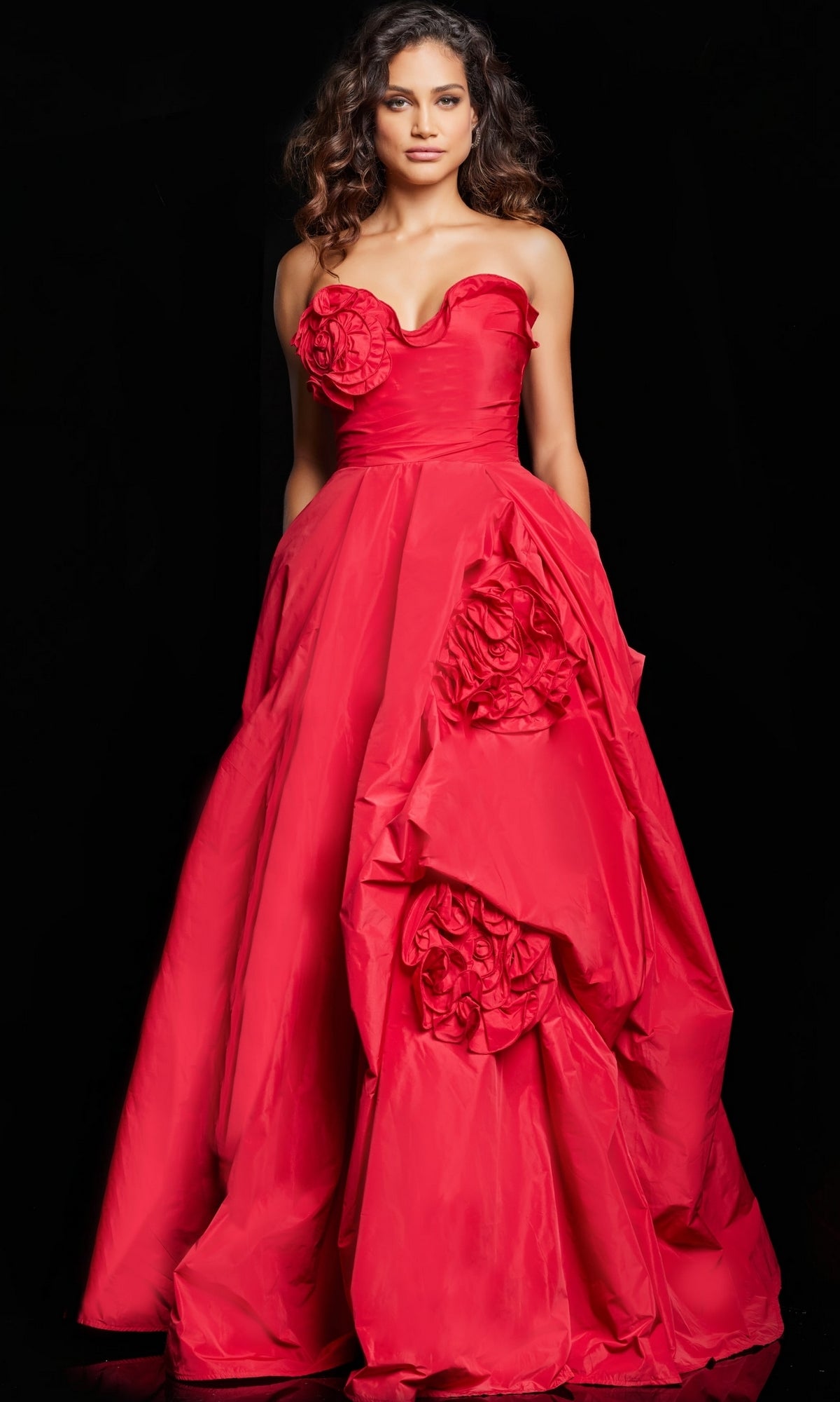 A woman stands against a black background wearing the Long Prom Dress 37266 by Jovani, a strapless, floor-length red ball gown adorned with large fabric roses on the bodice and skirt.