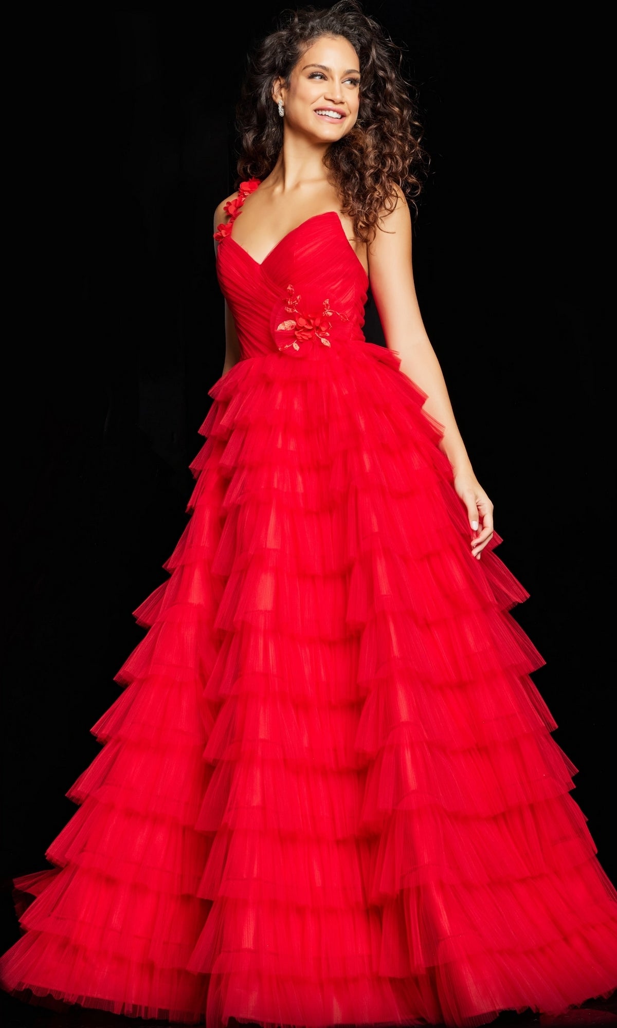 A woman stands against a black background wearing the Long Prom Dress 37274 by Jovani, a sleeveless, floor-length red ball gown with tiered tulle skirt and floral bodice detailing.
