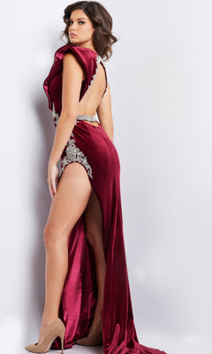 A woman wears the Long Prom Dress 37341 by Jovani, featuring deep red velvet, a high slit, open back, and crystal embellishments, paired with beige heels against a plain light background.