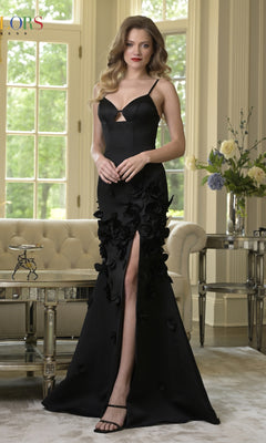 A woman in a black Cut-Out Long Floral Prom Dress (Colors Dress 3736) featuring a thigh-high slit stands in an elegant, well-lit living room with large windows and floral arrangements.