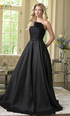A woman wearing the Colors Dress 3737, a strapless long A-line prom dress, stands in an elegant living room filled with light, featuring large windows and floral arrangements.
