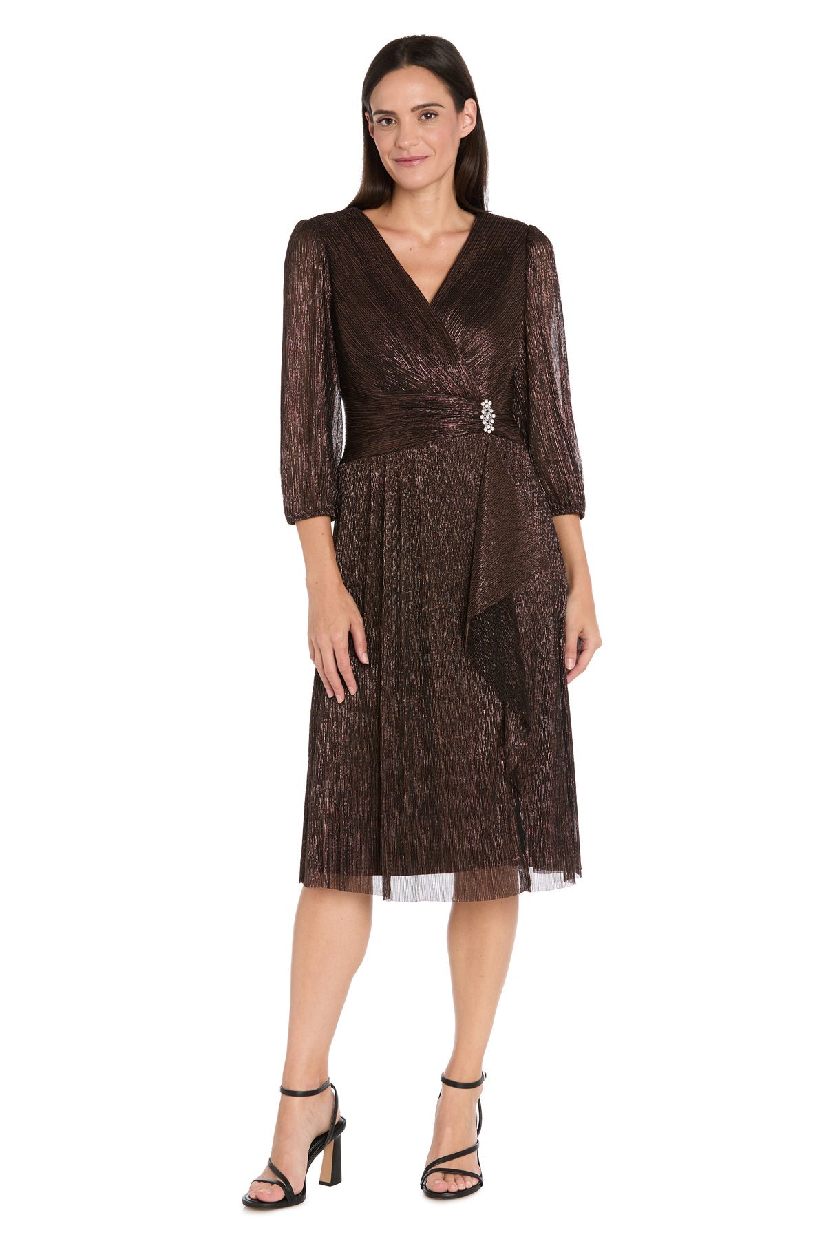 A woman models the R&M Richards 3741 Metallic Knee-Length Party Dress with long sleeves, V-neckline, pleated skirt, and black high-heeled sandals, standing against a white background.