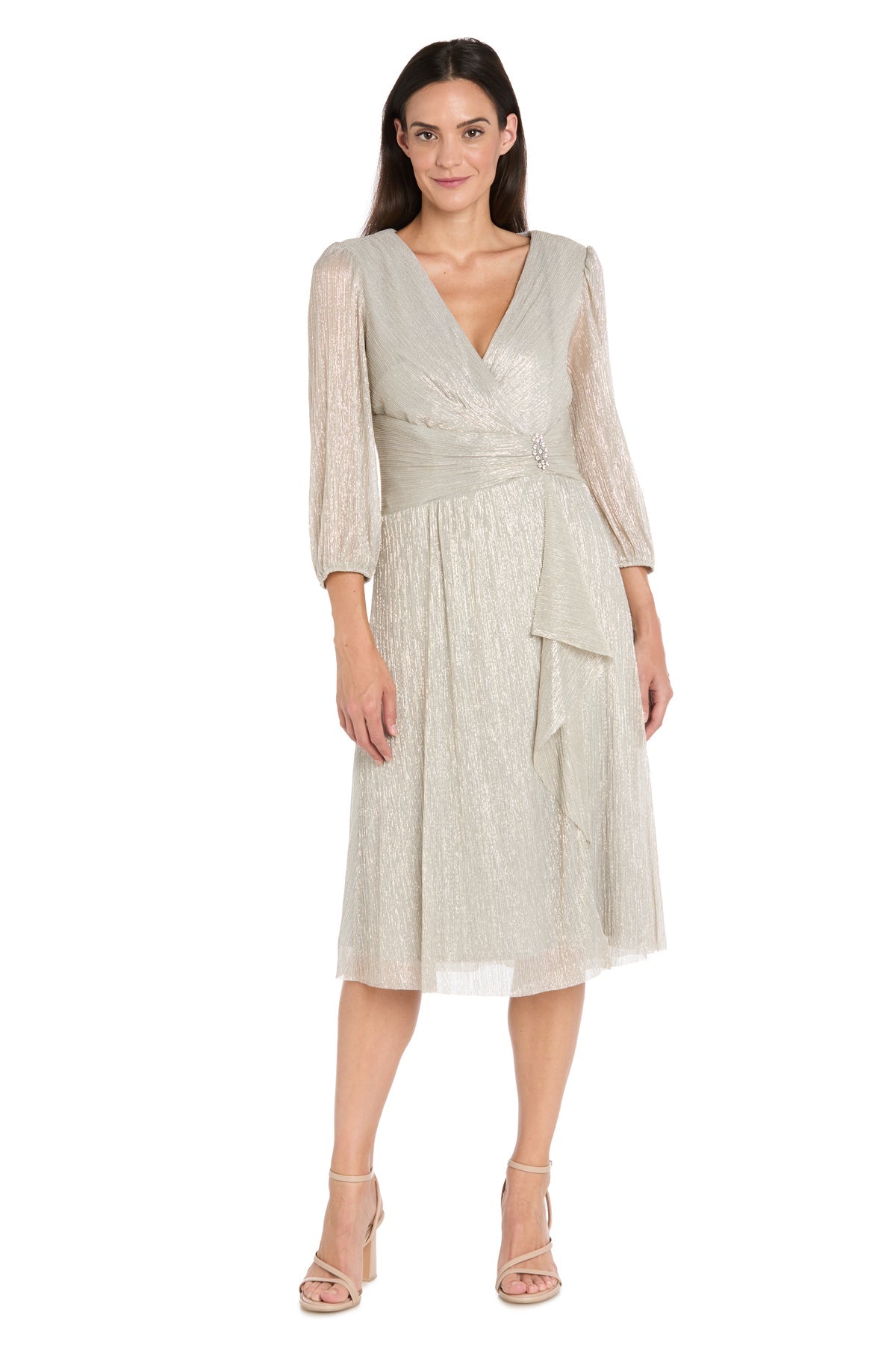 A woman wears the R&M Richards 3741 Metallic Knee-Length Party Dress—a light gold wrap design with sheer sleeves and a decorative waist brooch—elegantly paired with nude heeled sandals.