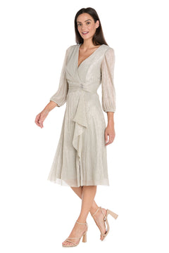 A woman models the R&M Richards 3741 Metallic Knee-Length Party Dress, featuring three-quarter sleeves and a decorative waist brooch, paired with nude heeled sandals against a plain white background.