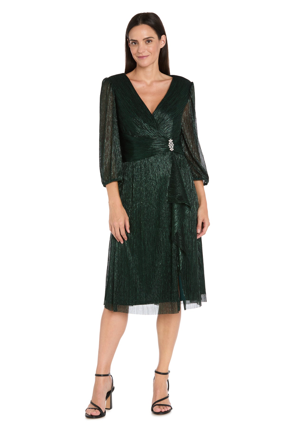 Woman in the R&M Richards 3741 Metallic Knee-Length Party Dress, featuring sheer long sleeves, V-neckline, gathered waist with jeweled brooch, and black heeled sandals, standing against a white background.