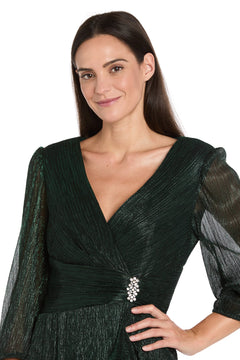 A woman with long dark hair wears the R&M Richards 3741 Metallic Knee-Length Party Dress in shimmery dark green with sheer sleeves and a jeweled brooch, standing against a white background.