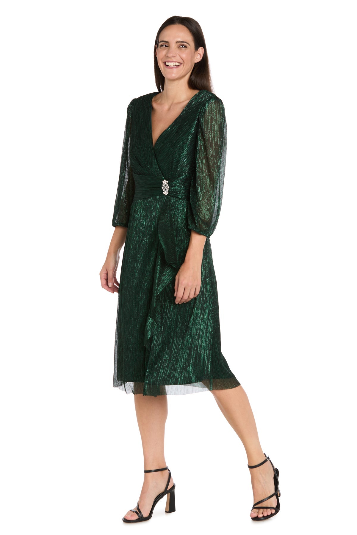 A woman wears the Metallic Knee-Length Party Dress: R&M Richards 3741, featuring shimmery green fabric, long sheer sleeves, waist embellishment, and black heeled sandals—a perfect semi-formal look.