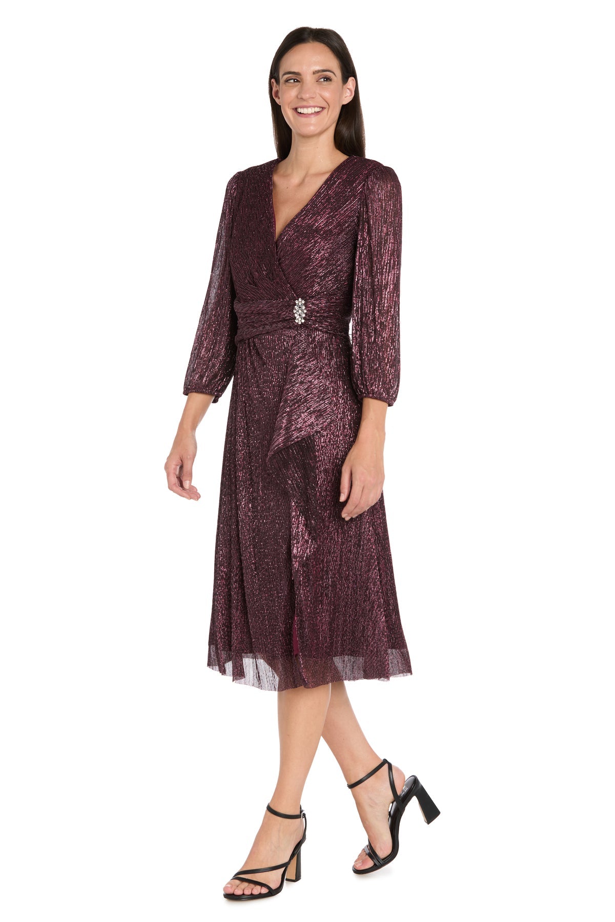 A woman in the R&M Richards 3741 Metallic Knee-Length Party Dress—a shimmery burgundy, long-sleeve wrap style with waist brooch—paired with black strappy heels.