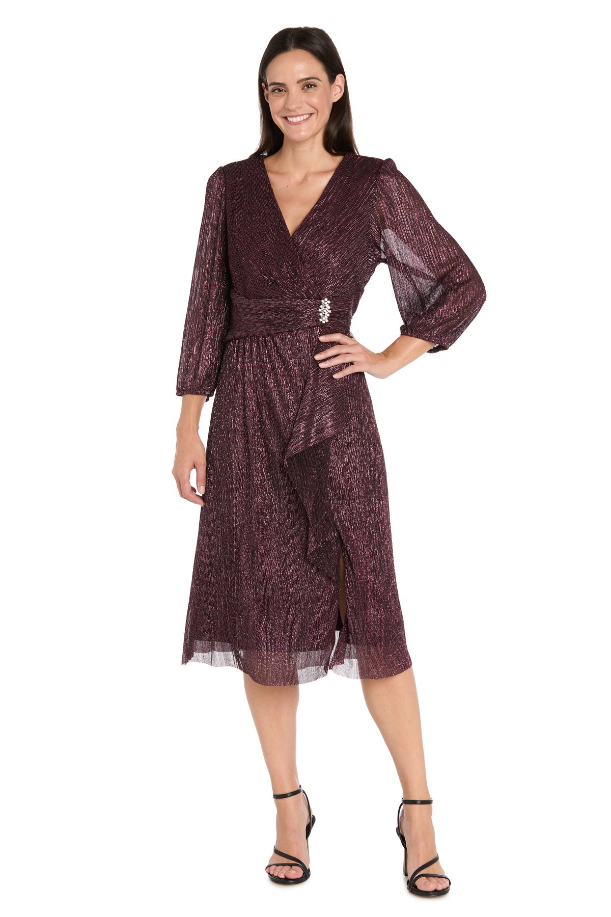 A woman in the R&M Richards 3741 Metallic Knee-Length Party Dress with long sleeves and a shimmery maroon finish, featuring a decorative waist brooch and black strappy heels, stands against a white background.