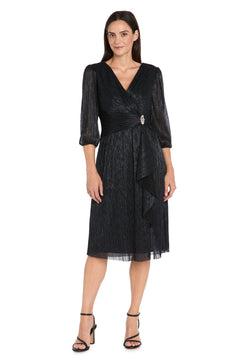 A woman models the R&M Richards 3741 Metallic Knee-Length Party Dress with 3/4 sleeves, V-neckline, waist brooch, and black ankle-strap heels against a white background.