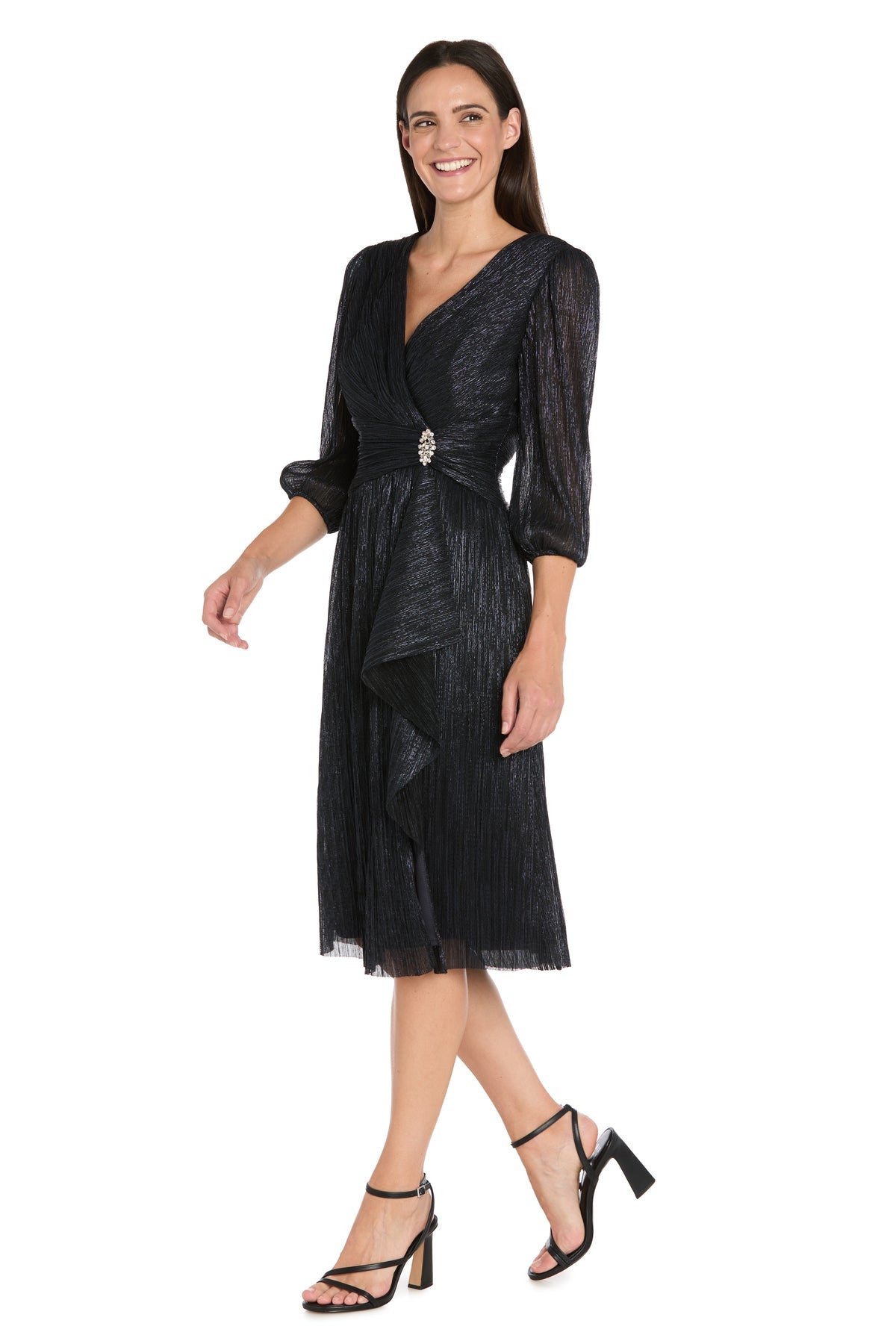 A woman stands smiling in the Metallic Knee-Length Party Dress: R&M Richards 3741, featuring a V-neck, three-quarter sleeves, waist brooch accent, and black heeled sandals.