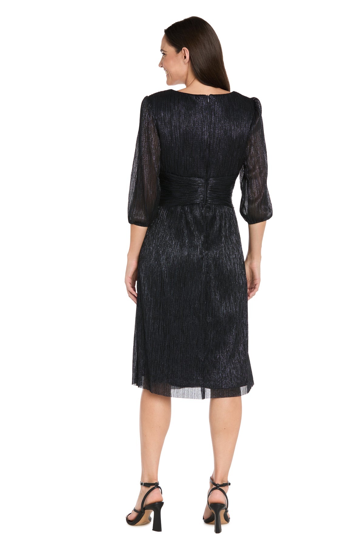 A woman stands facing away in the R&M Richards 3741 Metallic Knee-Length Party Dress with three-quarter sleeves and black heeled sandals. The shimmery black fabric enhances the elegance of this semi-formal look against a white background.