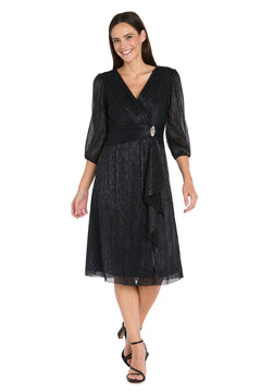A woman stands smiling in the R&M Richards 3741 Metallic Knee-Length Party Dress, featuring three-quarter sleeves and a decorative waist brooch.